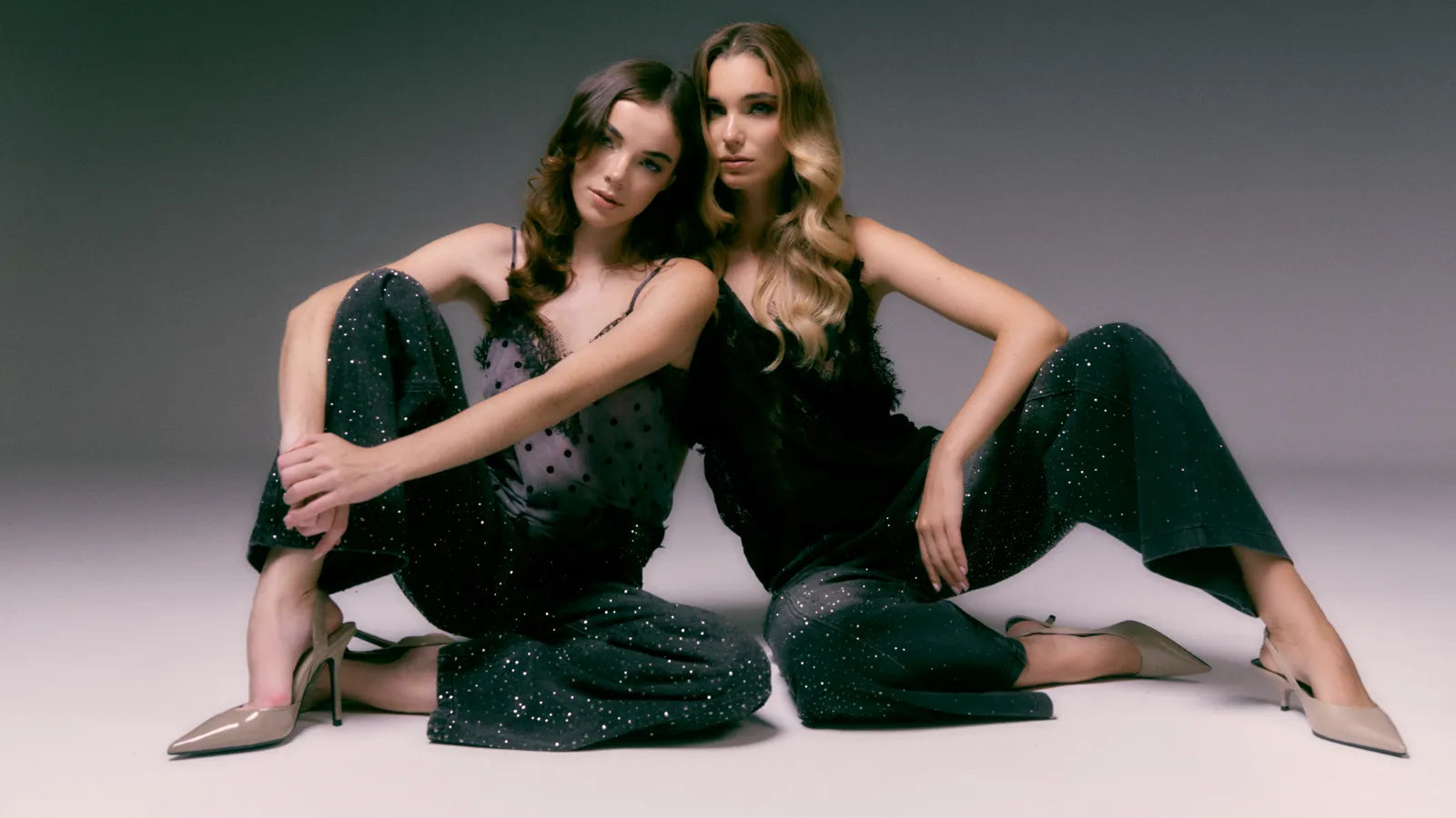 Two women in sparkly green dresses sitting on a gray background