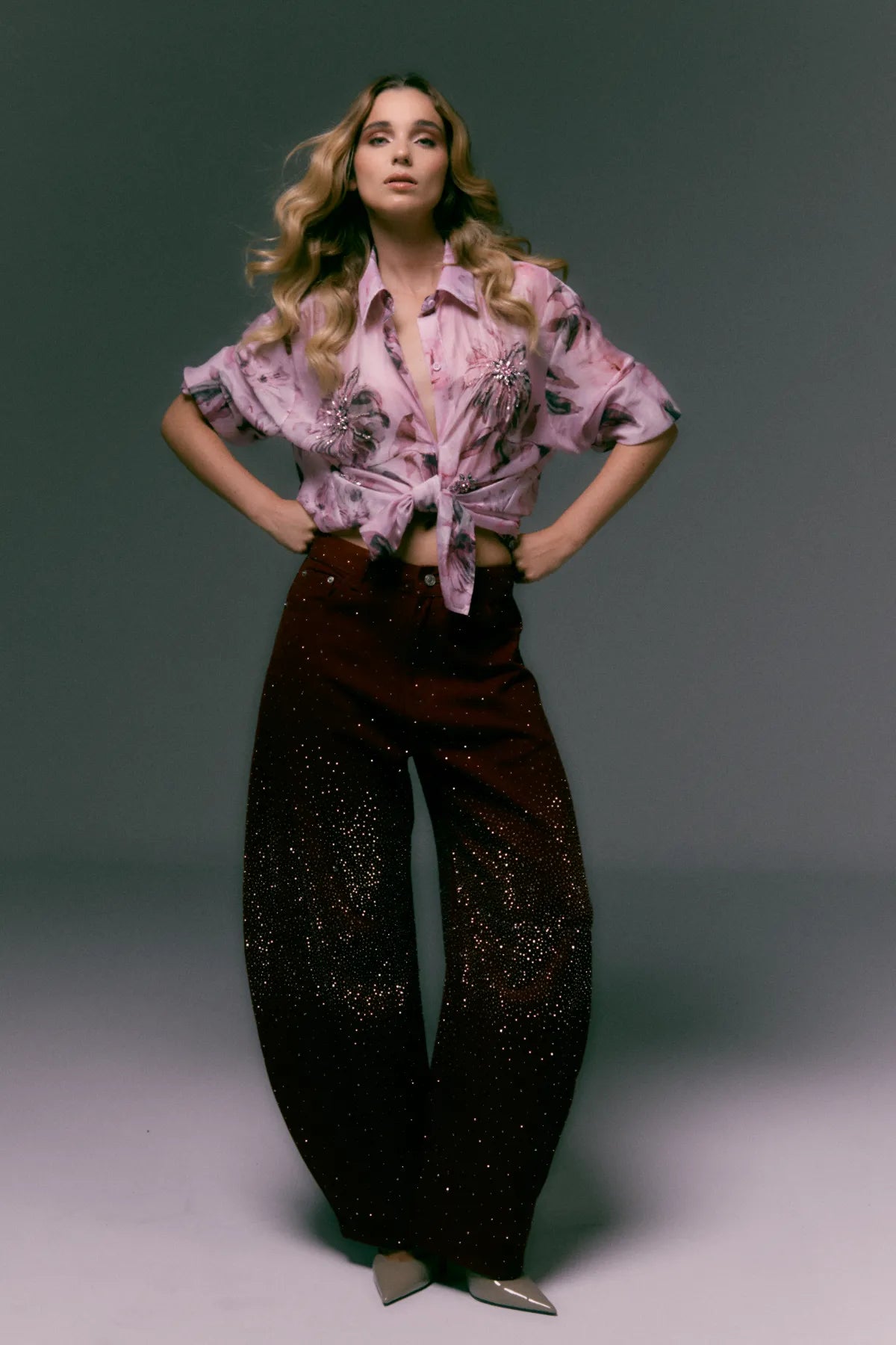 Woman wearing a patterned blouse and dark pants against a gray background